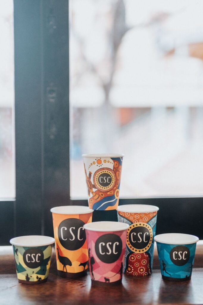 A stack of coffee cups sitting on a desk with an Aboriginal artwork on it and 'CSC' written in the middle.