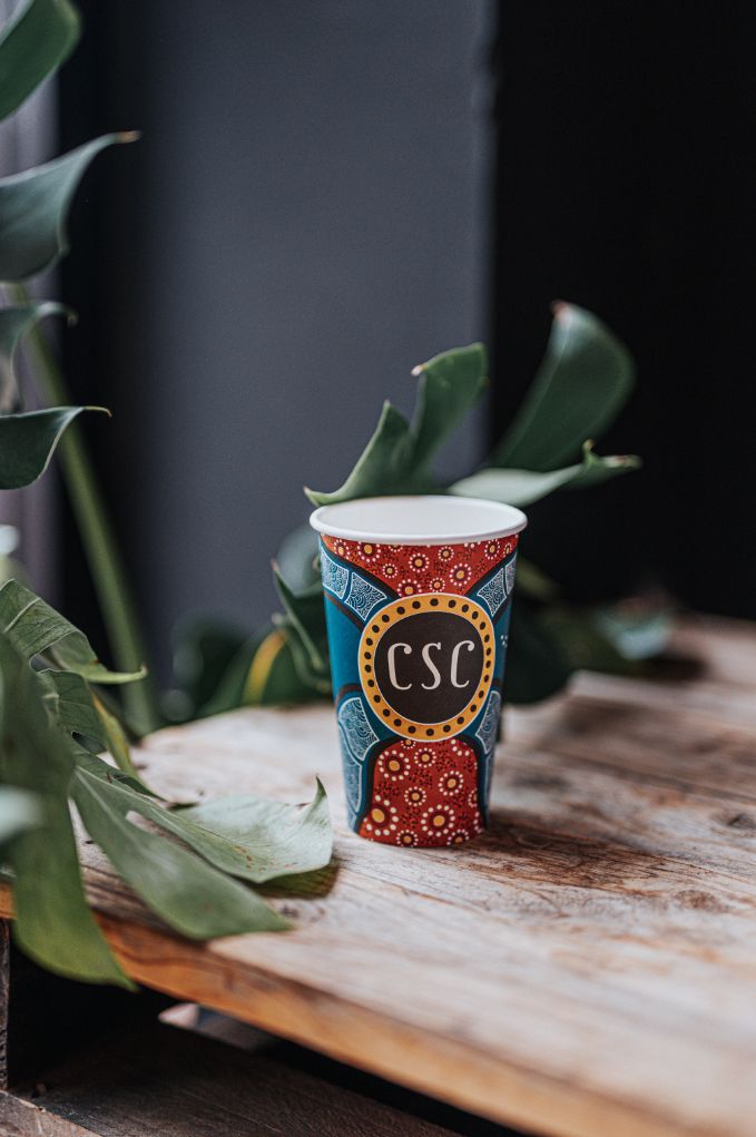 A coffee cup sitting on a desk with an Aboriginal artwork on it and 'CSC' written in the middle.