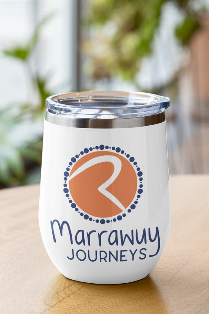 A small travel cup sitting on a table with an orange and dark blue circular logo saying 'Marrawuy Journeys".